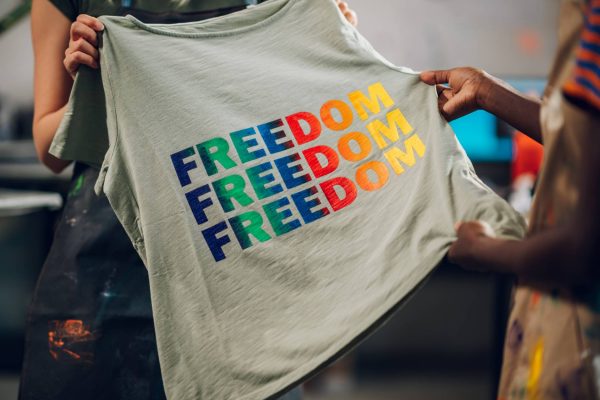 Two textile workers holding up a grey t-shirt with the word "freedom" printed three times on the shirt in bright, rainbow colors.