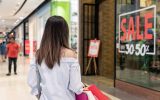 The back of a woman holding shopping bags who is looking at a storefront with "SALE, 30-50%" written on the glass.