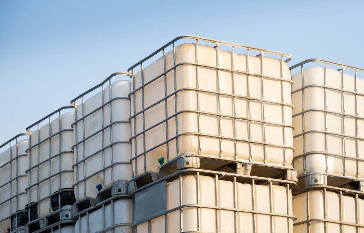 Rows of white plastic chemical tanks are stacked on top of each other. They are outside and the sky is blue.