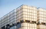 Rows of white plastic chemical tanks are stacked on top of each other. They are outside and the sky is blue.