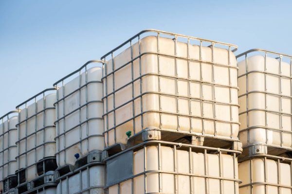 Rows of white plastic chemical tanks are stacked on top of each other. They are outside and the sky is blue.