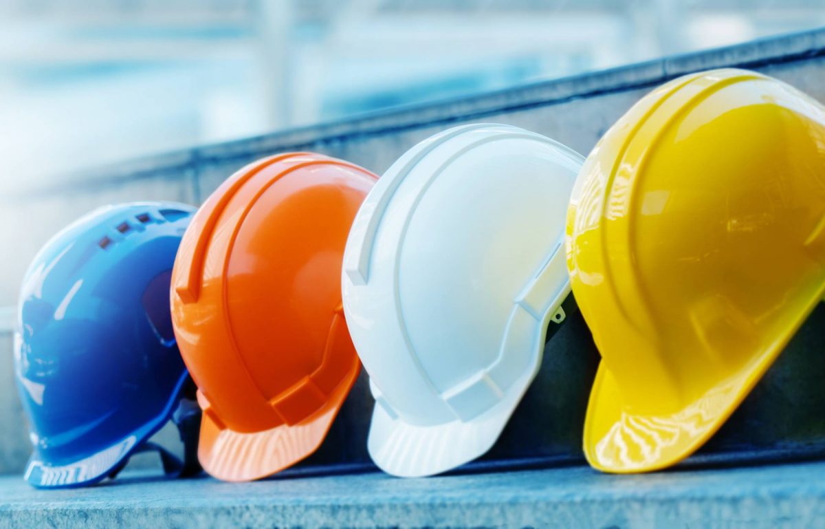 An orange, a yellow, a white, and a blue hard hat sit in a line on a concrete step. The background is blurry.