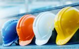 An orange, a yellow, a white, and a blue hard hat sit in a line on a concrete step. The background is blurry.