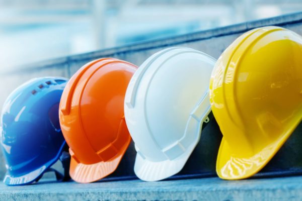 An orange, a yellow, a white, and a blue hard hat sit in a line on a concrete step. The background is blurry.