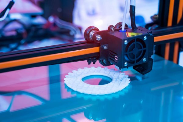 A large 3D printing machine with a small moving square is printing a white, round piece on top of a plastic base.