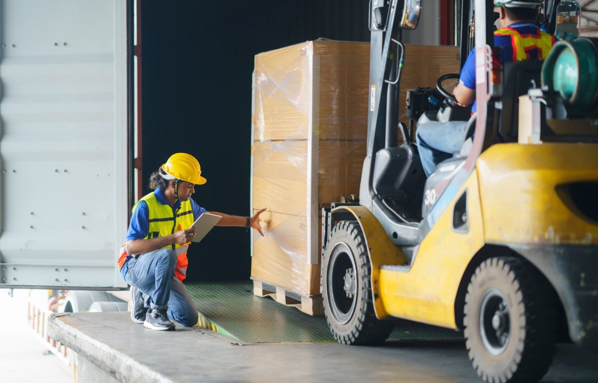 A person driving a forklift is driving a pallet of goods into a truck in a loading dock. Another employee wearing PPE is helping.