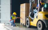 A person driving a forklift is driving a pallet of goods into a truck in a loading dock. Another employee wearing PPE is helping.