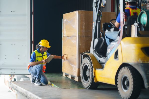 A person driving a forklift is driving a pallet of goods into a truck in a loading dock. Another employee wearing PPE is helping.