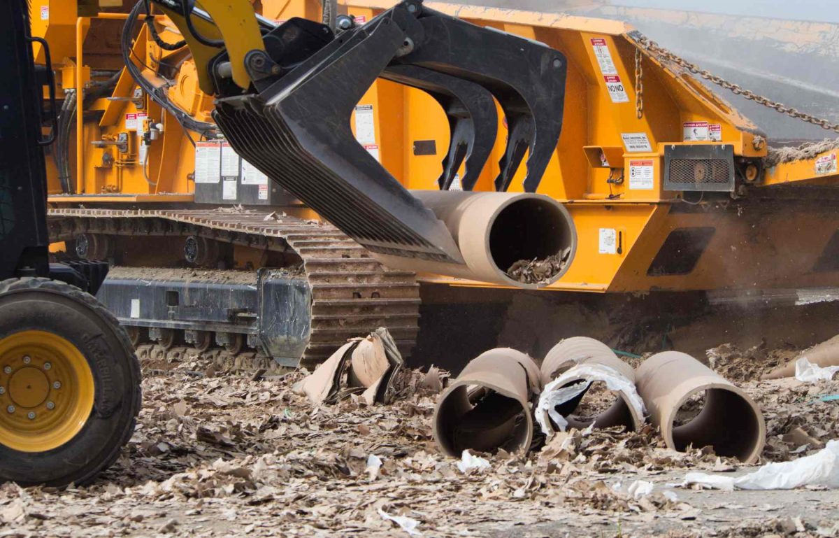 A yellow skid steer with a black grapple bucket is using the attachment to pick up a dirty pipe full of debris.