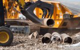 A yellow skid steer with a black grapple bucket is using the attachment to pick up a dirty pipe full of debris.