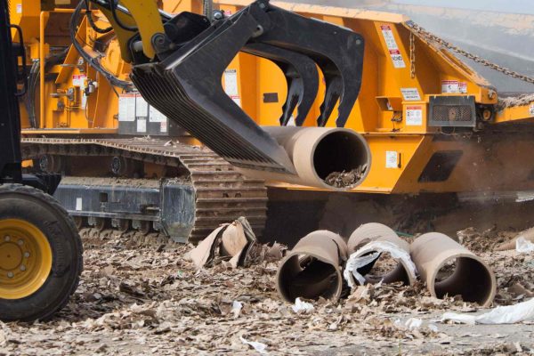 A yellow skid steer with a black grapple bucket is using the attachment to pick up a dirty pipe full of debris.