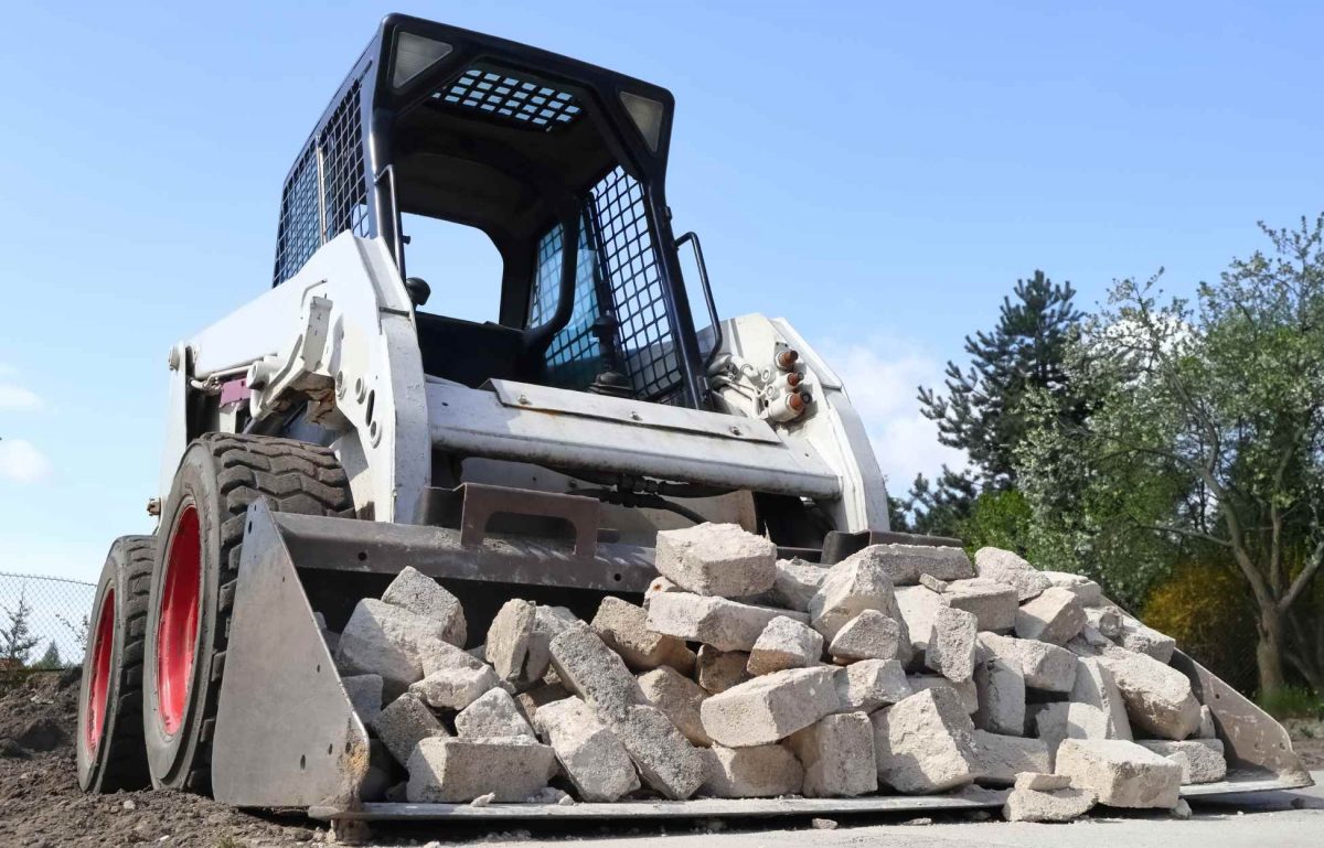 A black and white skid steer is moving a load of white bricks. The skid steer is driving on a dirt path surrounded by trees.