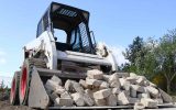 A black and white skid steer is moving a load of white bricks. The skid steer is driving on a dirt path surrounded by trees.