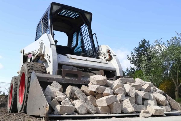 A black and white skid steer is moving a load of white bricks. The skid steer is driving on a dirt path surrounded by trees.