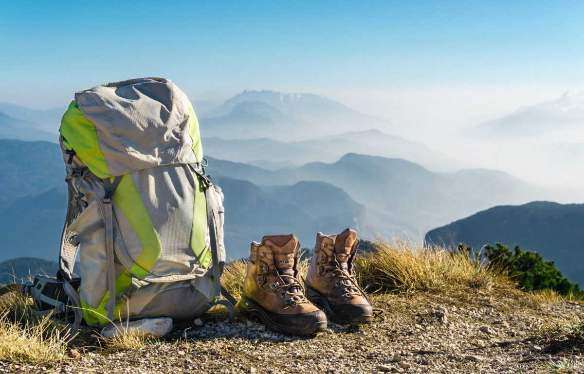 A backpack and a pair of hiking boots sitting on a trail with foggy mountain peaks and a blue sky in the background.