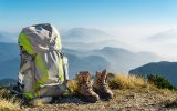 A backpack and a pair of hiking boots sitting on a trail with foggy mountain peaks and a blue sky in the background.