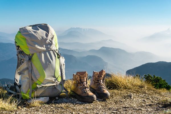 A backpack and a pair of hiking boots sitting on a trail with foggy mountain peaks and a blue sky in the background.