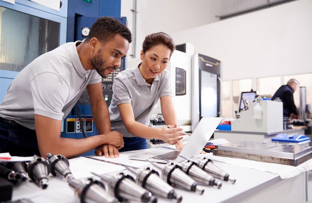 Two engineers looking at a piece of paper while leaning on a table full of multiple metallic products of the same type.