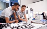 Two engineers looking at a piece of paper while leaning on a table full of multiple metallic products of the same type.