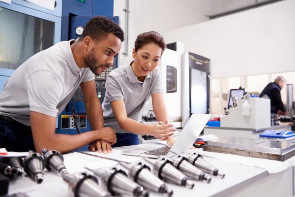 Two engineers looking at a piece of paper while leaning on a table full of multiple metallic products of the same type.