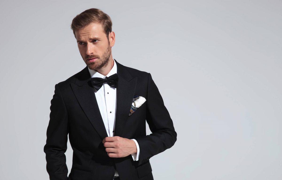 Young man posing while wearing a black tuxedo with a white shirt, a black bowtie, and a white handkerchief in front of a white wall.