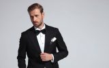 Young man posing while wearing a black tuxedo with a white shirt, a black bowtie, and a white handkerchief in front of a white wall.