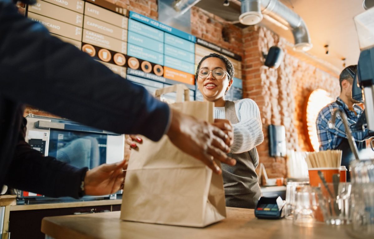 A woman in a white sweater and brown apron pushes a brown paper bag across the counter to a person in a blue coat.