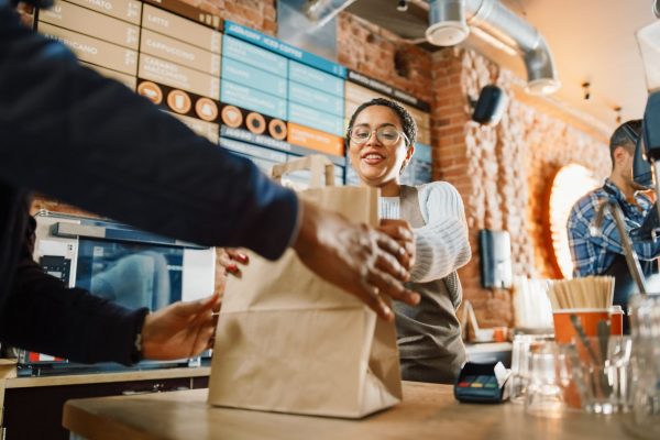 A woman in a white sweater and brown apron pushes a brown paper bag across the counter to a person in a blue coat.