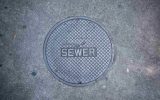 A large, gray manhole cover sits on a cracked asphalt road on a hole and it contains the word "sewer."