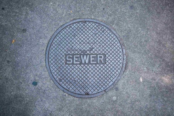 A large, gray manhole cover sits on a cracked asphalt road on a hole and it contains the word "sewer."