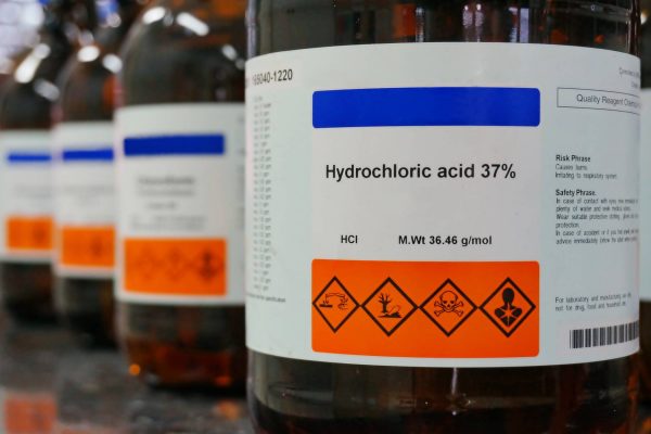 A close-up of a row of bottles with white labels on them. The front label says "Hydrochloric acid 37%" and has a hazard warning.