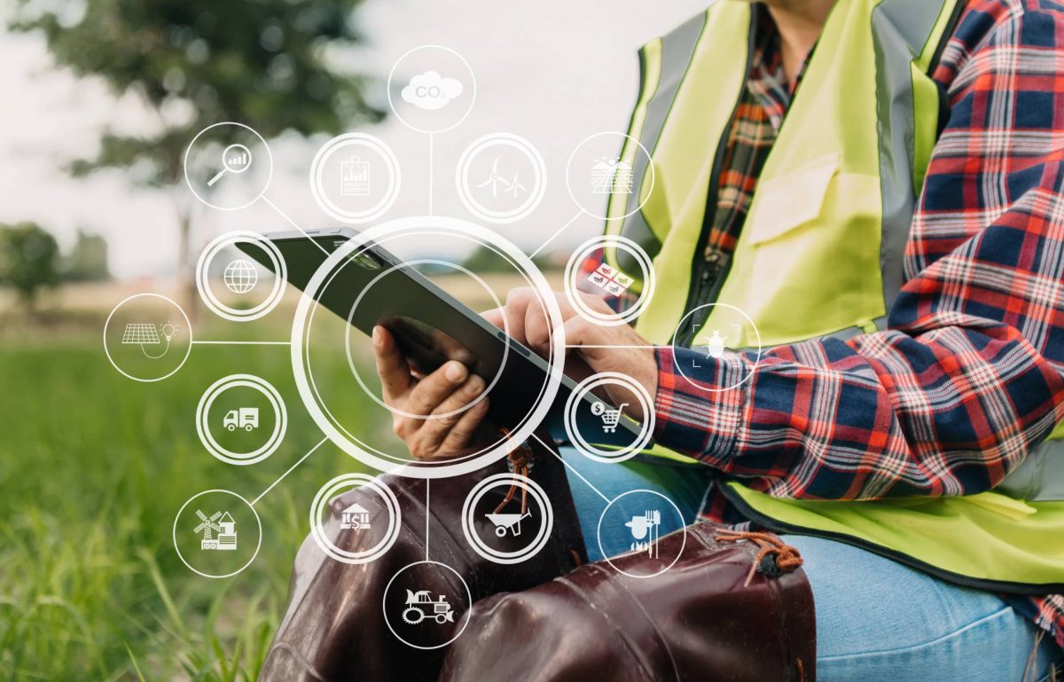 A farmer in a plaid shirt and neon vest sits in a field and holds a tablet with abstract data floating around it.