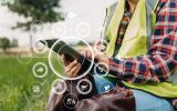 A farmer in a plaid shirt and neon vest sits in a field and holds a tablet with abstract data floating around it.