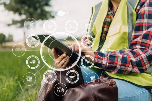 A farmer in a plaid shirt and neon vest sits in a field and holds a tablet with abstract data floating around it.