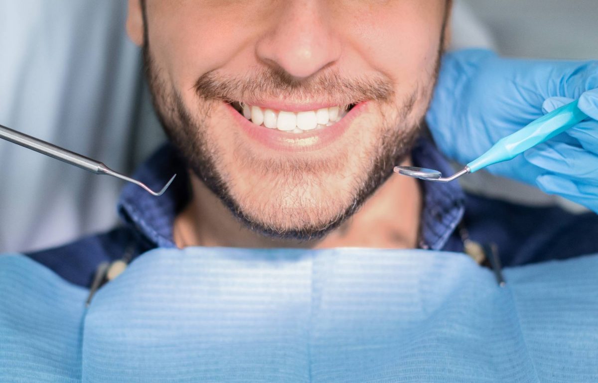 A man with a dental bib smiles, revealing a white, straight smile, as gloved hands with dental tools hover near his face.