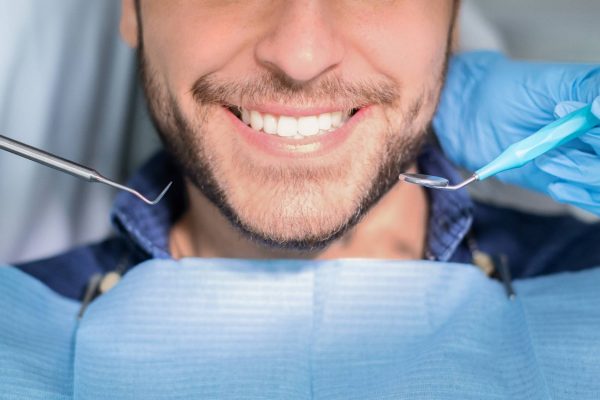 A man with a dental bib smiles, revealing a white, straight smile, as gloved hands with dental tools hover near his face.