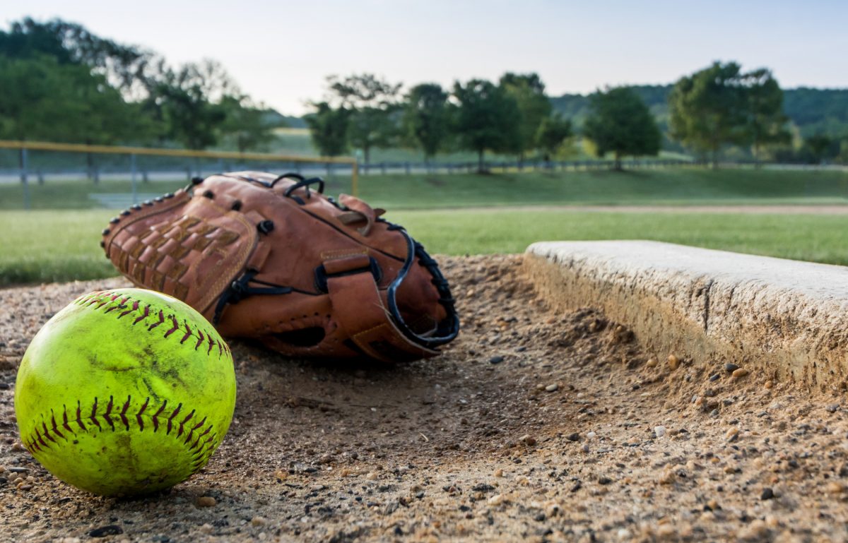 A softball and a glove sitting on the diamond. The pitcher's mound is next to the ball and glove and looks used.