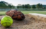 A softball and a glove sitting on the diamond. The pitcher's mound is next to the ball and glove and looks used.