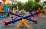 A vibrant, colorful powder-coated playground with a colorful seesaw in the foreground with trees behind it.