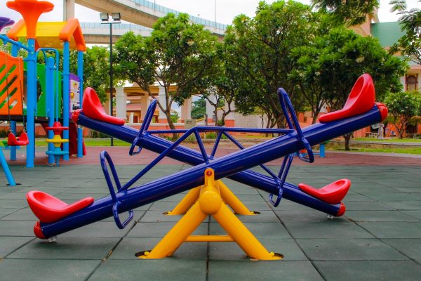 A vibrant, colorful powder-coated playground with a colorful seesaw in the foreground with trees behind it.