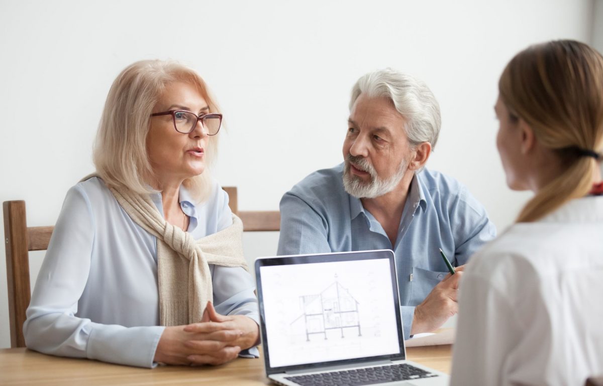 An older couple sits at a table discussing with a third person while a laptop displaying a house blueprint is open.