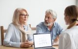 An older couple sits at a table discussing with a third person while a laptop displaying a house blueprint is open.