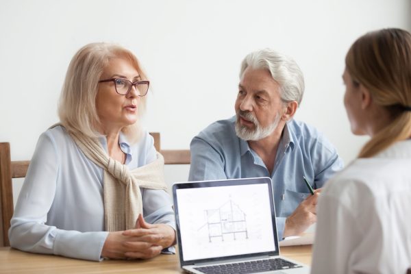 An older couple sits at a table discussing with a third person while a laptop displaying a house blueprint is open.