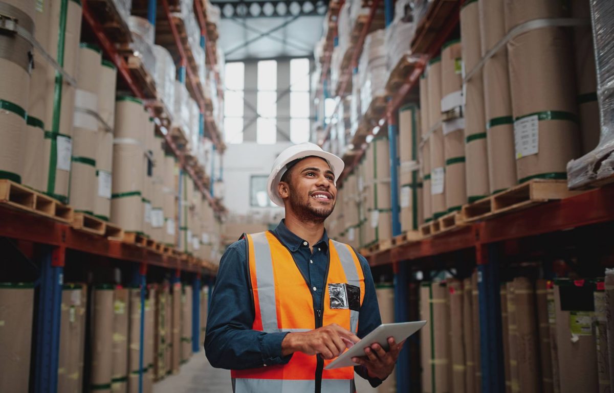 A warehouse worker wearing a hard hat and orange vest smiles as he taps on a tablet and walks down an inventory aisle.