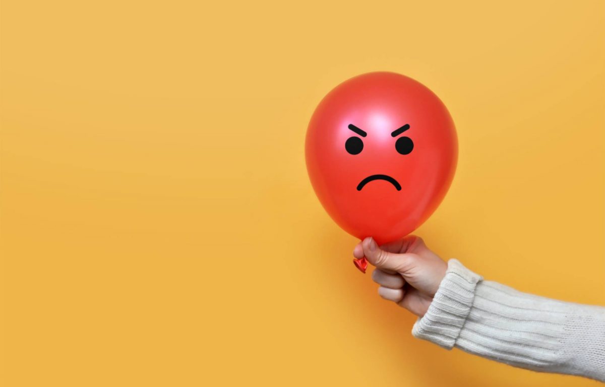 A person wearing a white sweater is holding a red balloon with an angry face. The background is orange.