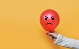 A person wearing a white sweater is holding a red balloon with an angry face. The background is orange.