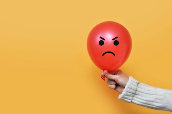 A person wearing a white sweater is holding a red balloon with an angry face. The background is orange.