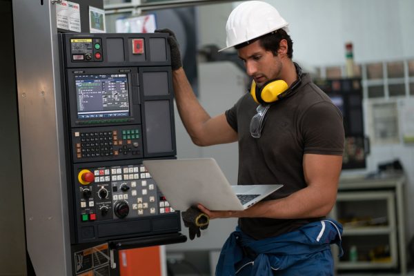 An engineer working on their laptop in a busy factory warehouse. He is standing next to a panel with many buttons.