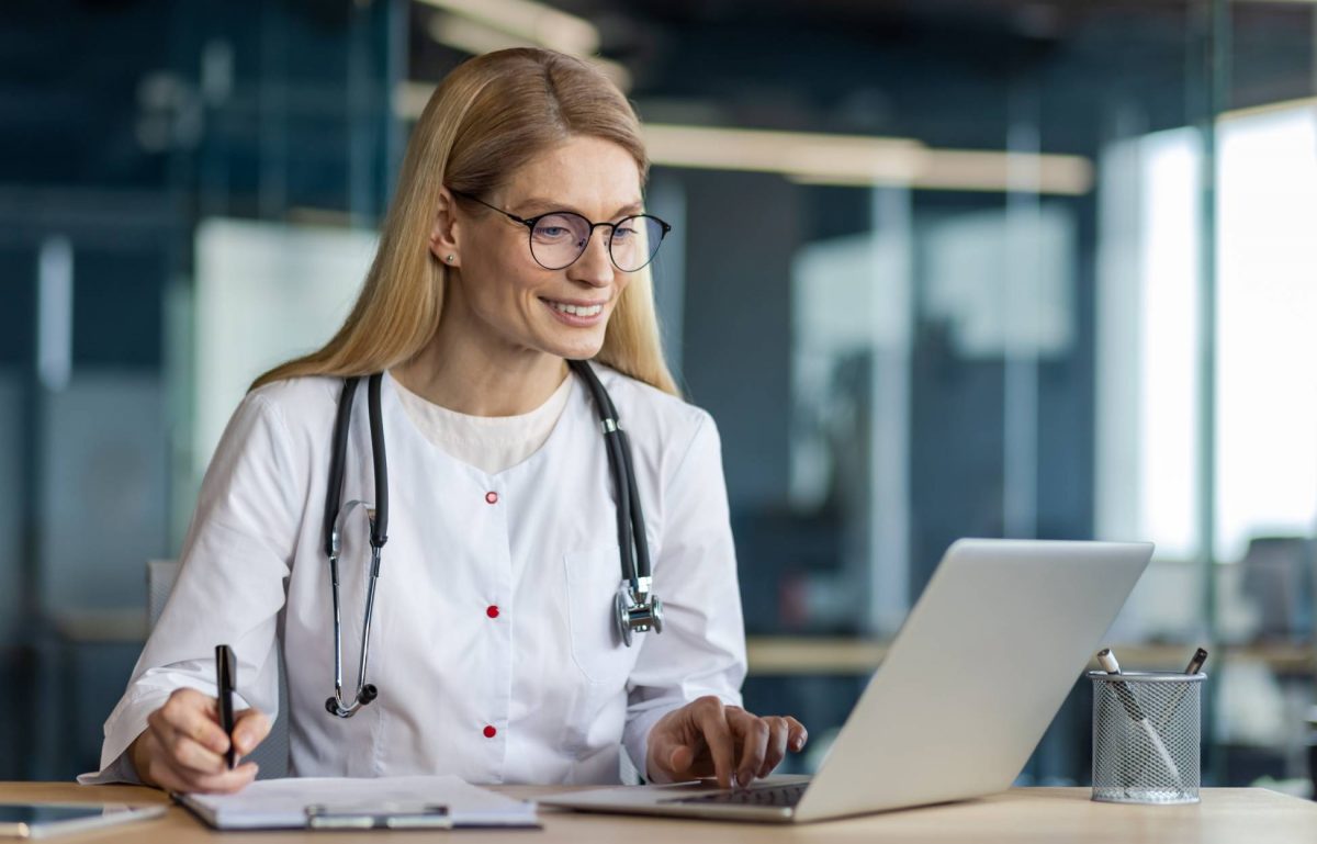 A medical professional wearing a stethoscope around her neck is using her laptop and writing something on her clipboard.