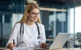 A medical professional wearing a stethoscope around her neck is using her laptop and writing something on her clipboard.